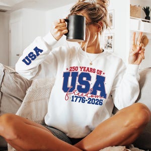 May include: White sweatshirt with the text "USA" on the left sleeve and a larger design on the front that reads "250 Years of USA Freedom 1776-2026" in blue and red. The person is holding a black mug.