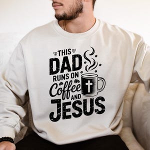 May include: Cream-colored sweatshirt with a black graphic that reads "THIS DAD RUNS ON Coffee AND JESUS." The design includes a coffee mug with a cross and steam rising from it. The sweatshirt has long sleeves and a crew neck.