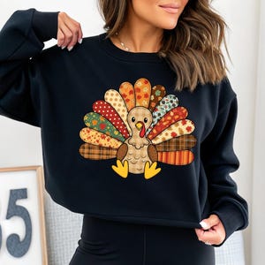 May include: Black sweatshirt featuring a colorful cartoon turkey design. The turkey has a patchwork-style tail with various patterns and colors, including orange, red, and brown. The design is centered on the front of the sweatshirt, perfect for Thanksgiving.
