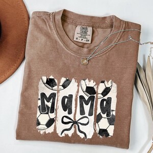 Puede incluir: Una camiseta marr&oacute;n Comfort Colors con la palabra "Mama" en letras negras, acentuada con balones de f&uacute;tbol y un lazo. Un collar plateado con un colgante est&aacute; drapeado sobre la camisa. La camisa es de talla peque&ntilde;a.