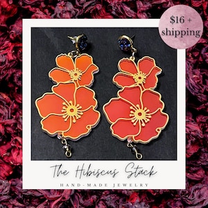 Puede incluir: Un par de pendientes florales hechos a mano. Cada pendiente presenta dos formas de flores naranjas y rojas en capas con centros y contornos dorados. Los pendientes tienen pequeños detalles en azul oscuro en la parte superior y cuentas colgantes. El texto "The Hibiscus Stack" está en la parte inferior.