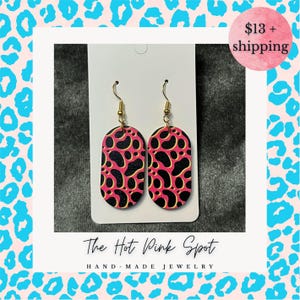 May include: A pair of oval-shaped earrings with a pink background and black spots. The earrings have gold-colored hooks. The text "The Hot Pink Spot" and "HAND-MADE JEWELRY" are at the bottom of the image.