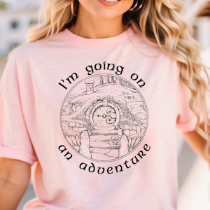 以下が含まれることがあります： ホビットの穴の黒いグラフィックと「冒険に出かける」というテキストが書かれたピンクのTシャツです。