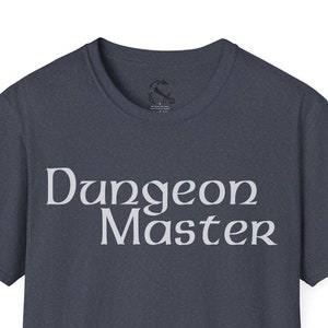 May include: A navy blue t-shirt with the words "Dungeon Master" printed in a stylized, light gray font. The shirt has short sleeves and a crew neck. A small logo is visible near the neckline.