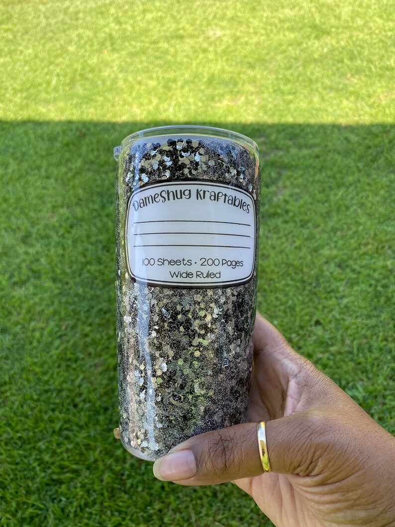 May include: A black and gold glitter tumbler with a white label that reads "DameShug Kraftables 100 Sheets - 200 Pages Wide Ruled".