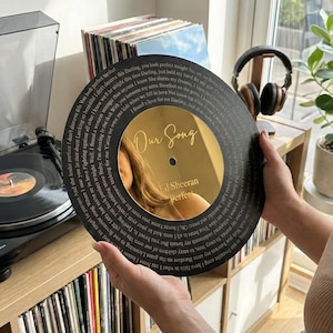 May include: A black vinyl record with the words "Our Song" and the artist's name, Ed Sheeran, engraved on a gold-coloured centre. The record is held in front of a record player and a collection of vinyl records.