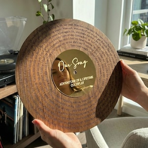 Könnte beinhalten: Eine Holzplatte mit eingravierten Texten und einem goldfarbenen Mittelteil. In der Mitte der Platte stehen die Worte "Our Song" und "Adventure of a Lifetime Coldplay". Die Platte wird von einer Person gehalten.