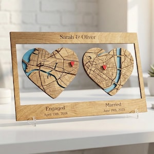May include: Wooden framed artwork featuring two heart-shaped map cutouts with red pin markers. The frame includes the names "Sarah & Oliver" and the dates "Engaged April 13th, 2018" and "Married June 19th, 2023". The maps have blue accents.