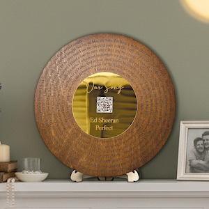 May include: A circular decorative item with a wooden appearance, featuring the lyrics of Ed Sheeran's "Perfect" around the edge. The centre has a gold-toned reflective surface with the words "Our Song", a QR code, and the song title and artist.