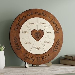 Può includere: Puzzle familiare in legno circolare con bordo marrone scuro e pezzi chiari. Il testo "together we make a family" è inciso sul bordo. Nomi individuali e la data "July 17, 2023" sono incisi. Un pezzo a forma di cuore presenta un albero.