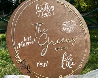 May include: A round wooden wedding sign with the words "Wedding Time," "Save the Date," and "Love of my Life." The sign also includes the names "The Greens" and "EST 2024." The sign is decorated with gold and white accents.
