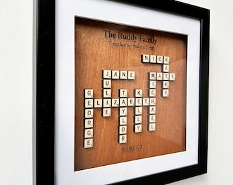 May include: A framed wood plaque with the words "The Ruddy Family" and "Together we make a family" at the top. The plaque features a Scrabble-style crossword puzzle with the names "Jane", "Nick", "Matt", and "Elizabeth" spelled out using letter tiles.