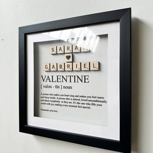 May include: A framed print with the names "Sara" and "Gabriel" spelled out with wooden letter tiles. The print also includes the word "Valentine" defined as "a person who makes you heart sing and makes you feel warm and fuzzy inside. A person who is adored, loved unconditionally and taken completely as they are. It's the one who fills your world with joy, making every moment feel special. Someone you love."