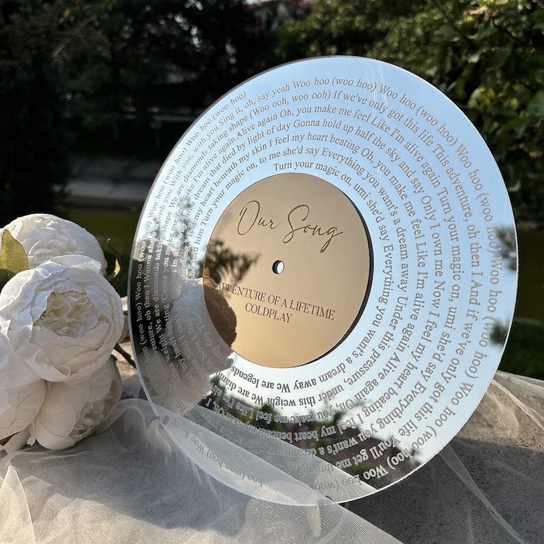 以下が含まれることがあります： 「Our Song」と「Adventure of a Lifetime」の文字が金色で刻まれた、反射する円形の装飾品。外縁には歌詞が記されています。白い花とシアーな生地と一緒に展示されています。
