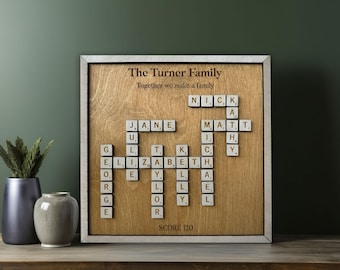 May include: A wooden frame with a light brown background and a white border. The frame contains a scrabble tile crossword puzzle with the names "Nick", "Jane", "Matt", "Elizabeth", and "George" spelled out. The text "The Turner Family" and "Together we make a family" is at the top of the frame. The text "Score 120" is at the bottom of the frame.