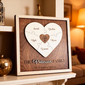 Può includere: Targa di famiglia in legno con un design a puzzle a forma di cuore. I pezzi del puzzle, di colore chiaro, hanno nomi incisi. Il centro del cuore presenta un disegno ad albero. La targa è incorniciata con un bordo in legno chiaro e include il cognome e una citazione.