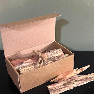 May include: A brown cardboard box containing several rectangular pieces of aromatic cedar wood tied together with a piece of natural jute twine. One additional piece of cedar wood with visible bark and reddish wood grain rests outside the box.