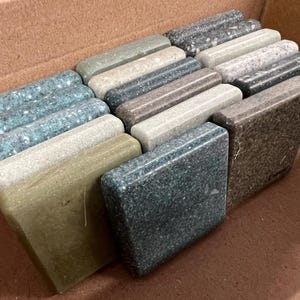 May include: A collection of rectangular stone samples in various colors, including teal, gray, and brown, with speckled patterns. The samples are arranged in a cardboard box, showcasing different textures and hues for design selection.