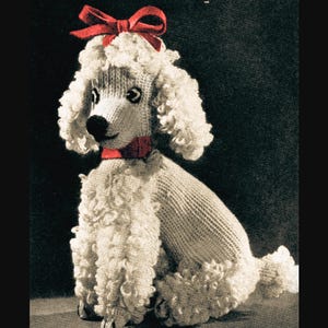 Könnte beinhalten: Ein gestrickter weißer Pudelhund mit lockigem Fell, einer roten Schleife auf dem Kopf und einem roten Halsband. Der Hund sitzt aufrecht, mit sichtbaren Vorderpfoten. Der Hintergrund ist dunkel.