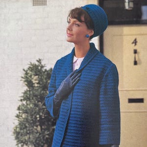May include: A woman wearing a blue crochet coat, matching hat, and gloves. The coat has a textured pattern and a collar. The woman is also wearing a pink collared shirt and blue earrings. The background includes a white wall, a green bush, and a door.