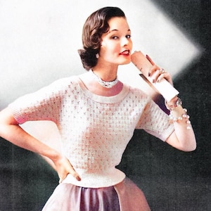 May include: A woman wearing a white knitted sweater with a delicate floral pattern. She is holding a white rolled-up paper in her right hand and has a pearl necklace and bracelet on.