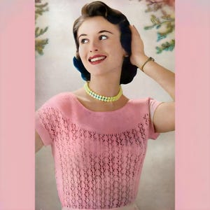 Könnte beinhalten: Ein rosa gestrickter Pullover mit einem zarten Spitzenmuster und kurzen Ärmeln. Der Pullover hat einen runden Ausschnitt und wird von einer Frau mit einer hellgelben und blauen Perlenkette getragen.