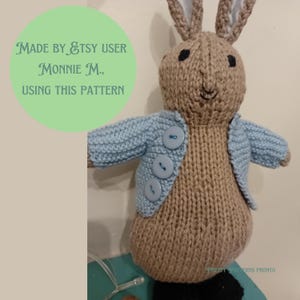 May include: A knitted brown bunny wearing a light blue cardigan with three blue buttons. The bunny is standing on a black surface. The text "MADE BY ETSY USER MONNIE M., USING THIS PATTERN" is visible in the image.
