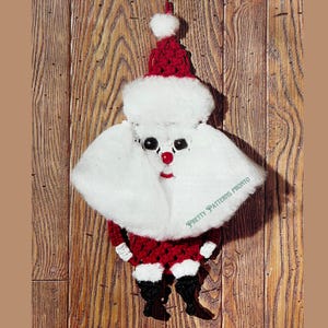 May include: A handmade Christmas decoration shaped like Father Christmas. It has a red crocheted hat and suit, a white fluffy beard and moustache, and black boots. The text "Pretty Patterns Pronto" is visible.