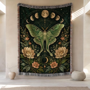 May include: A woven throw blanket featuring a large green moth centered against a dark background. The design includes a lunar cycle arch at the top, floral arrangements on both sides, and a crescent moon at the bottom. The edges have fringe.