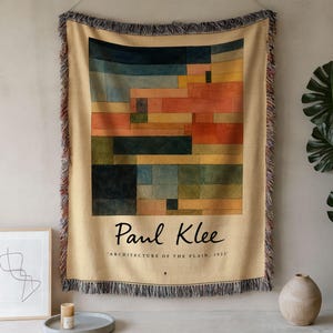 May include: A woven throw blanket featuring a rectangular abstract composition of colored blocks in shades of blue, orange, yellow, and green. The text Paul Klee and Architecture of the Plain, 1923 is printed below the art with fringed edges.