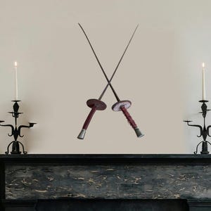 May include: Two antique fencing swords crossed in an X shape against a white wall. The swords have wooden handles and metal blades. The swords are displayed above a black marble fireplace mantel.