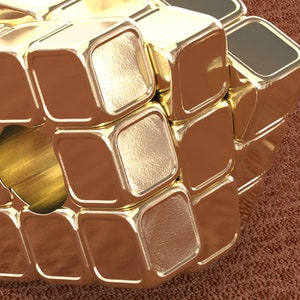 May include: A gold-colored metal cube puzzle with a hole in the center. The cube is made up of smaller squares that can be rotated.