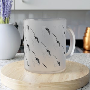 Puede incluir: Una taza blanca helada con un diseño en blanco y negro de pájaros estilizados. La taza tiene un asa y está sobre una superficie de madera.