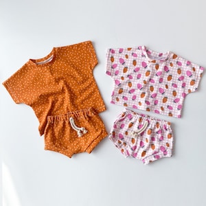May include: Two sets of children's clothing. One set is orange with a speckled pattern and the other is pink with a strawberry pattern. Both sets include a short-sleeved shirt and shorts.