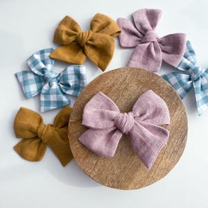 May include: A set of five fabric hair bows in various colors and patterns. The bows are made of a soft, textured fabric and are tied with a simple knot. The bows are arranged on a wooden surface.