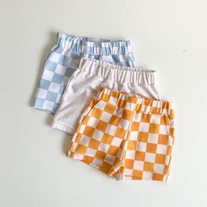 May include: Three pairs of checkered shorts in different colours: light blue, beige, and orange. The shorts have an elastic waistband and are made of a soft, comfortable fabric.