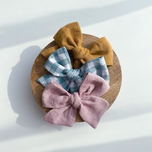 May include: Three fabric hair bows on a wooden surface. One bow is a solid mustard yellow, one is a blue and white gingham pattern, and one is a solid dusty rose.