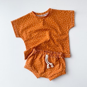 May include: A two-piece outfit for a baby or toddler, featuring a short-sleeved t-shirt and matching shorts. Both pieces are a burnt orange color with a white confetti print.