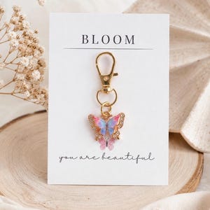 May include: A gold-toned metal keychain featuring a butterfly charm with pink, blue, and purple enamel wings accented by small clear crystals. The charm is attached to a white display card printed with the text BLOOM and you are beautiful.
