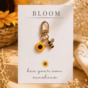 May include: A gold-toned metal keyring featuring a yellow sunflower charm and a black and yellow bee charm with clear crystals. The keyring is attached to a white display card printed with the text BLOOM and bee your own sunshine.