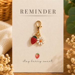 Pode incluir: Um pingente de metal dourado com fecho lagosta, apresentando um morango vermelho e uma margarida branca com centro laranja. O pingente está preso a um cartão de exibição creme com o texto REMINDER no topo e stay berry sweet na parte inferior.