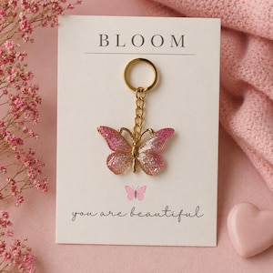 Könnte beinhalten: Ein goldfarbener Schmetterling-Schlüsselanhänger mit rosa und goldfarbenen Glitzerflügeln wird auf einer weißen Karte präsentiert. Die Karte trägt das Wort "BLOOM" oben und den Satz "you are beautiful" unten. Ein rosa Herz und rosa Blumen sind im Hintergrund.