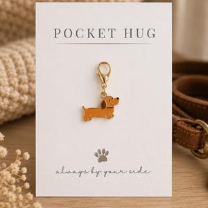 May include: A gold-toned metal charm shaped like a brown dachshund with a lobster clasp, attached to a white display card. The card features the text POCKET HUG at the top, a small grey paw print icon, and the phrase always by your side below.