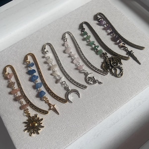 May include: A set of eight silver and gold bookmark charms with various gemstone and crystal accents. The charms feature a crescent moon, a star, a dragon, and a sun.