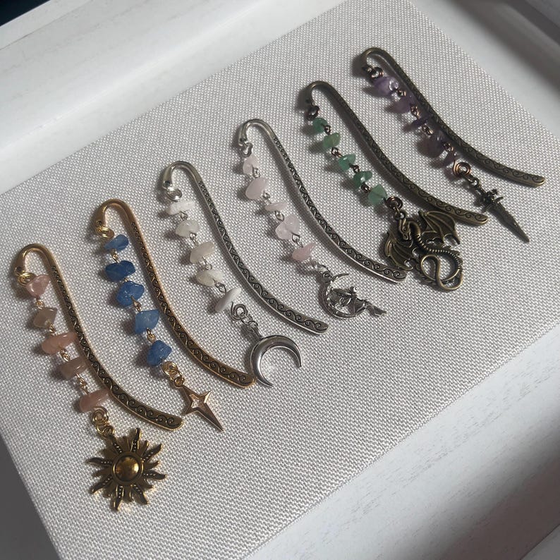 May include: A set of eight silver and gold bookmark charms with various gemstone and crystal accents. The charms feature a crescent moon, a star, a dragon, and a sun.