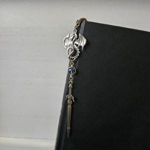 May include: Silver dragon bookmark with a sword charm and a blue iridescent bead. The bookmark is attached to a black book.