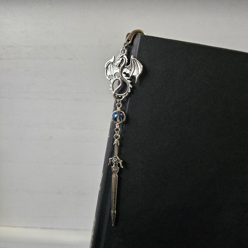 May include: Silver dragon bookmark with a sword charm and a blue iridescent bead. The bookmark is attached to a black book.