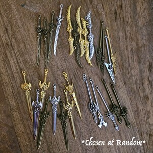May include: A collection of 18 silver and gold sword charms. The charms are arranged in two rows on a wooden surface. The charms are all different shapes and sizes, and some have intricate details. *Chosen at Random*