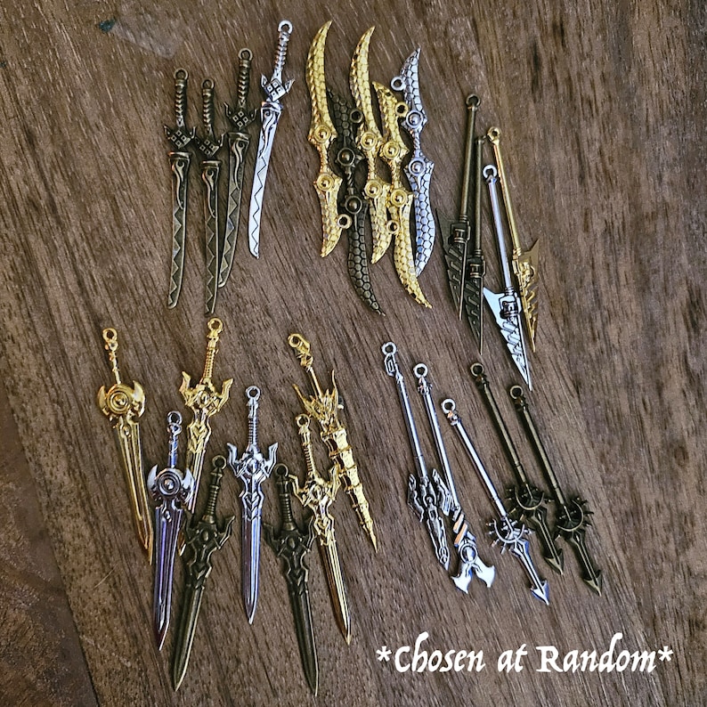 May include: A collection of 18 silver and gold sword charms. The charms are arranged in two rows on a wooden surface. The charms are all different shapes and sizes, and some have intricate details. *Chosen at Random*