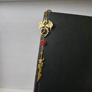 May include: Gold bookmark featuring a dragon design with a red gemstone and a sword pendant.